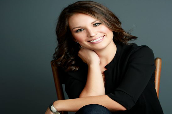 Portrait picture of a beautiful smiling woman with long dark brown hair, wearing a black blouse and dark jeans, looking directly at the camera with her right arm up to cheek supporting her titled head, and happy with her Ultherapy treatment at Premier Laser Center in San Jose, Costa Rica.